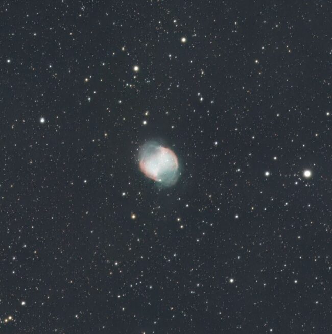 Dumbbell-shaped cloud of reddish and green nebulosity over a background of distant stars.