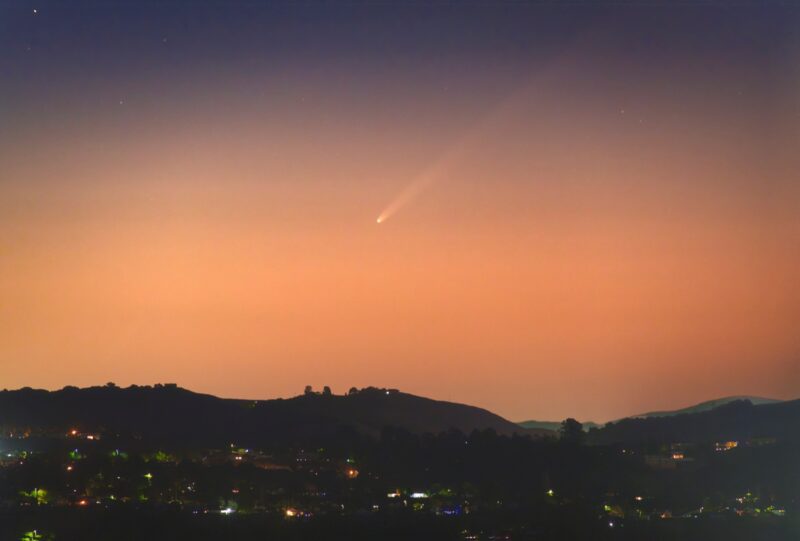 Orangish sky and dark landscape with some lights and comet above with long tail.