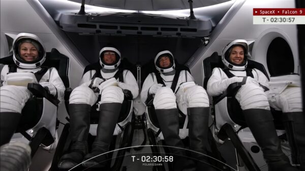 Four astronauts in spacesuits with open helmets, lined up in 4 seats in a crew capsule, smiling.