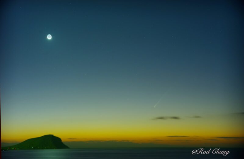 Moon at upper left with comet at lower right over the sea.