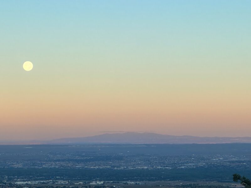 A colorful dawn sky with a bright white moon at left over a landscape with mountain.