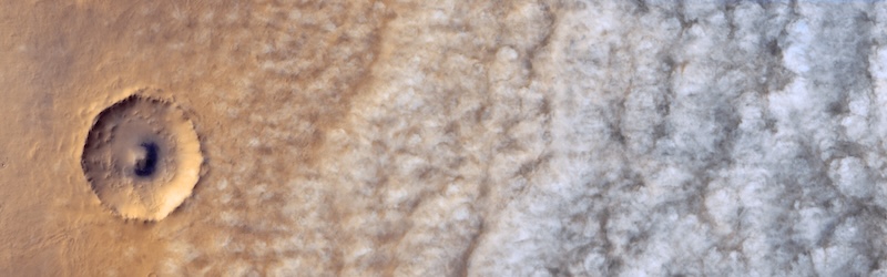 Crater in reddish terrain on the left and formation of fluffy white cloud lines on the right, seen from above.