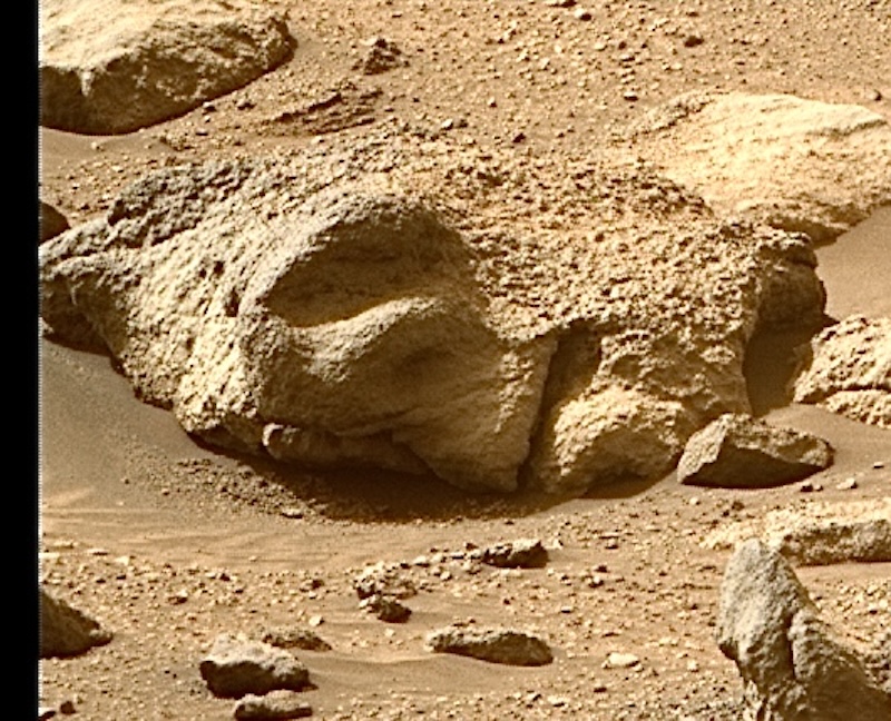 Oddly-shaped rock in reddish terrain that kind of looks like a human face lying sideways.