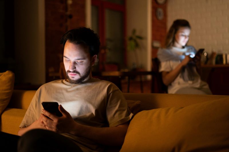 A man and woman in a darkened house looking at their lit smart phones.