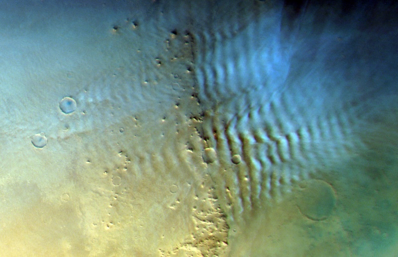 Bluish-white ripple-like clouds seen from above over reddish, cratered terrain.