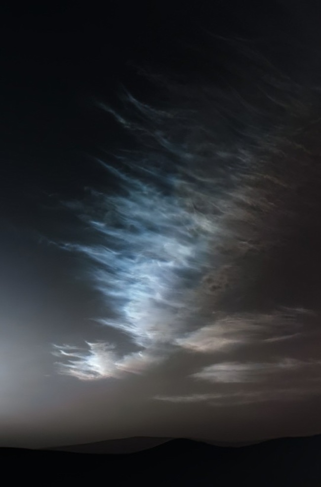 Wispy bluish clouds shining in night sky over distant hills.