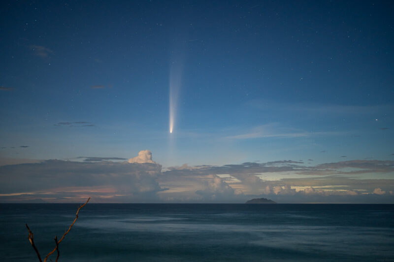 Comet above sea and clouds, with a big tail pointing upward and 2nd tail pointing downwards to the horizon.