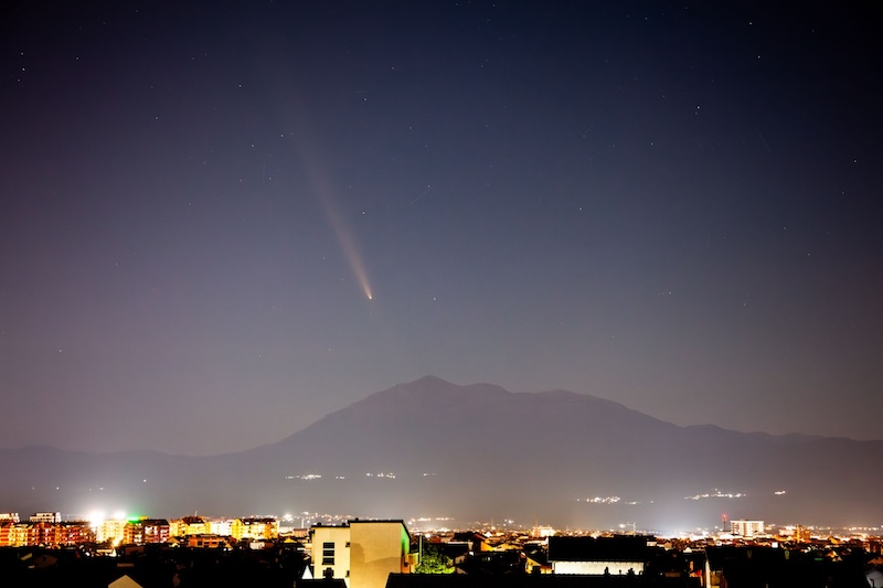 Long-tailed comet over a distant mountain and foreground city lights.