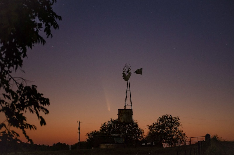 Twilight sky with trees in foreground and small comet in the sky next to an old-fashioned Texas windmill.