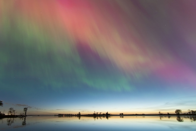 Green and red auroras in an early twilight sky above a lake.