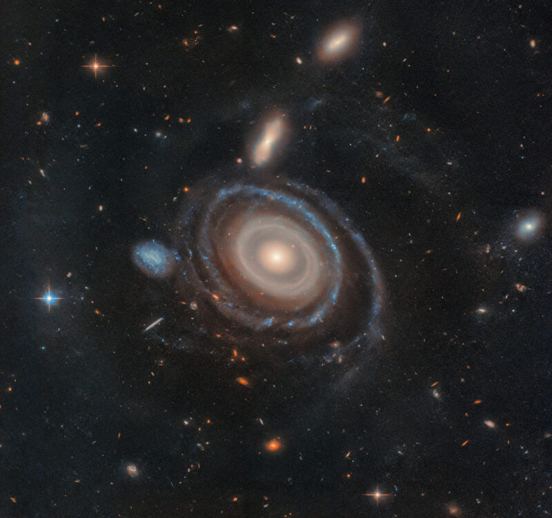 A galaxy with 9 rings in cream and blue tones and smaller galaxies in the background.