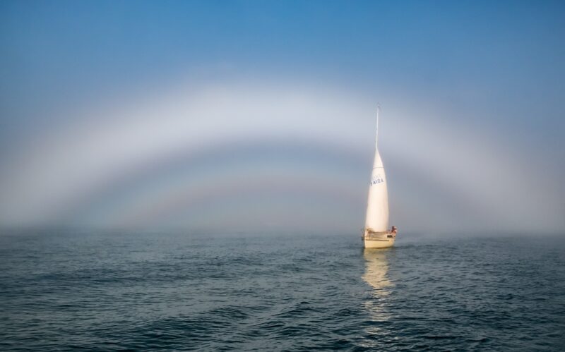 A white rainbow with a colorful, dimmer rainbow inside, plus the sea and a sailboat.