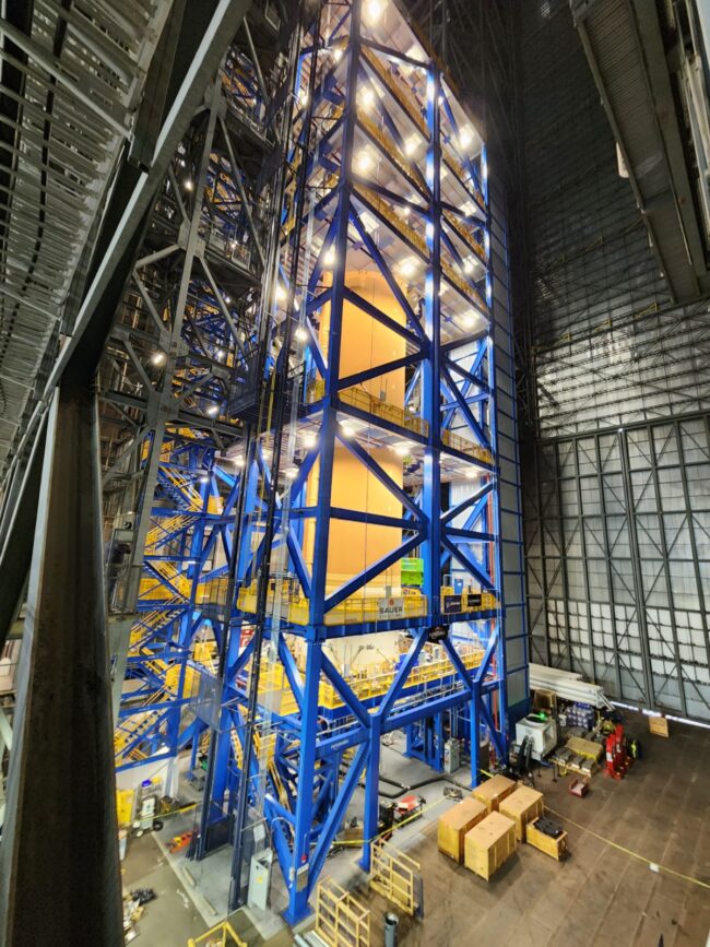Tall, square, open structure of blue girders surrounding a vertical orange cylinder.