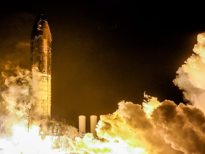 Night scene: a tall, metallic cylindrical rocket stands above billowing flame and smoke.