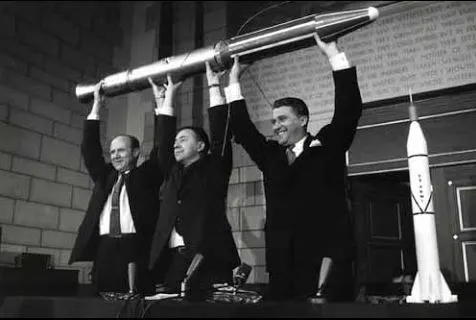 Three middle aged white men in suits hold a long model rocket above their heads, smiling.