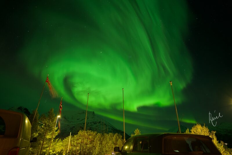 Bright, glowing swirls of curtain-like green light in the night sky above a parking lot.