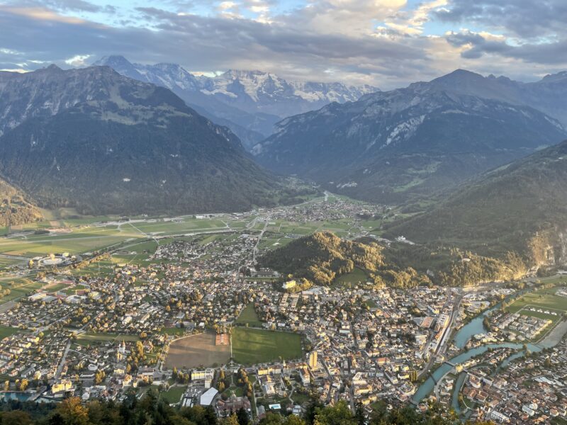 A city nestled between snow-capped mountains with a large treed hill.