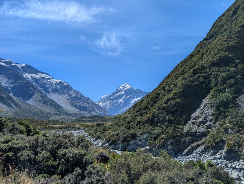 A tall snowy mountain peak in the distance below a blue sky, with a steep, grassy valley leading from it towards the viewer, who is on the valley floor.