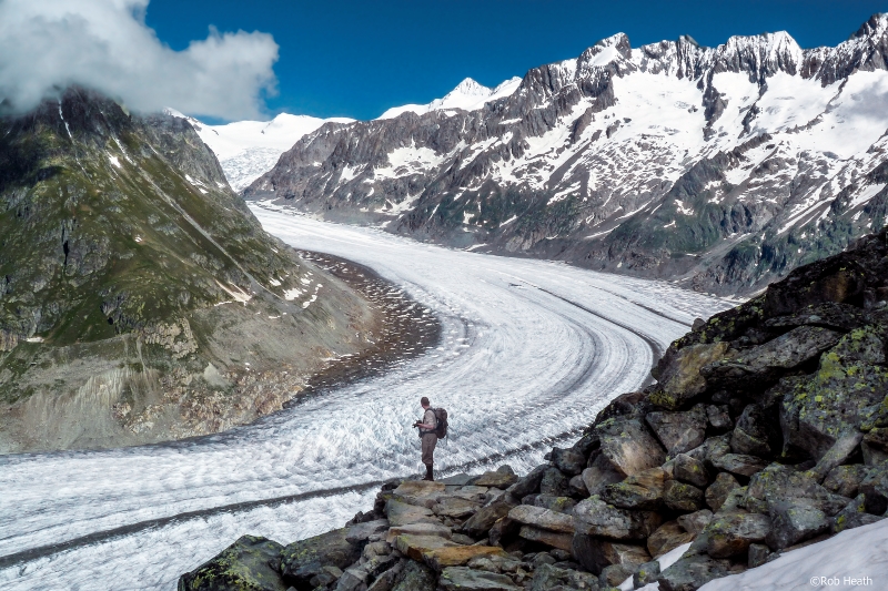 A wide icy stretch curves sharply through a rocky valley, with snow-covered peaks on the right and a man in hiking gear in the foreground.