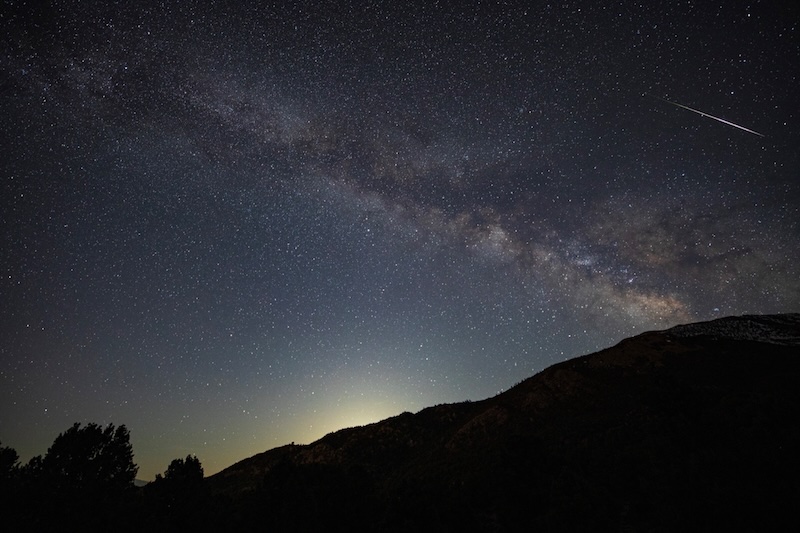 Fuzzy, glowing band of the Milky Way over a hill, with a short bright streak near it.