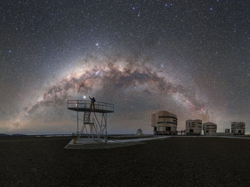 The Milky Way arching through a dark sky with observatory buildings and a man on a structure reaching upward.