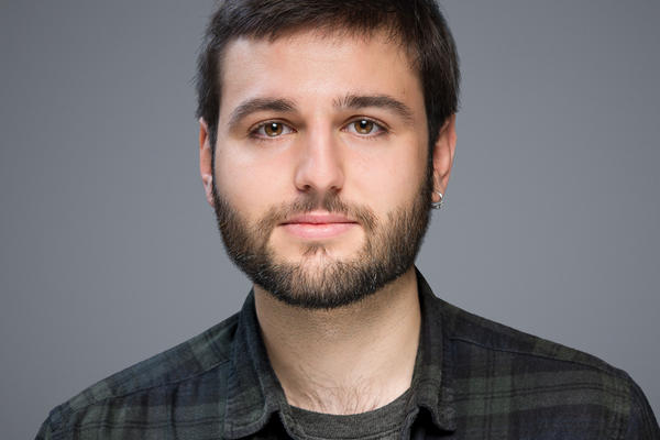 Young man with dark hair and facial hair with a smile in his eyes looking at the camera.