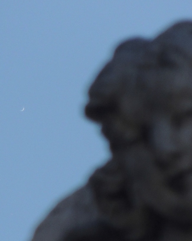 Tiny, thin crescent on a blue sky with out-of-focus part of a statue in the foreground.