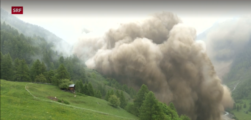 A huge, billowing, dirty cloud rolling down a forested hillside among mountains.
