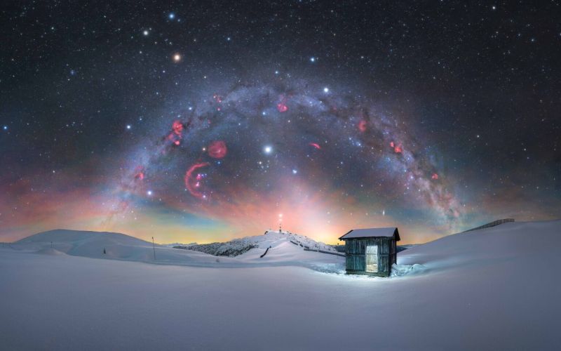 Snowy landscape and hut with arching Milky Way behind and scattered gas clouds along it in red.