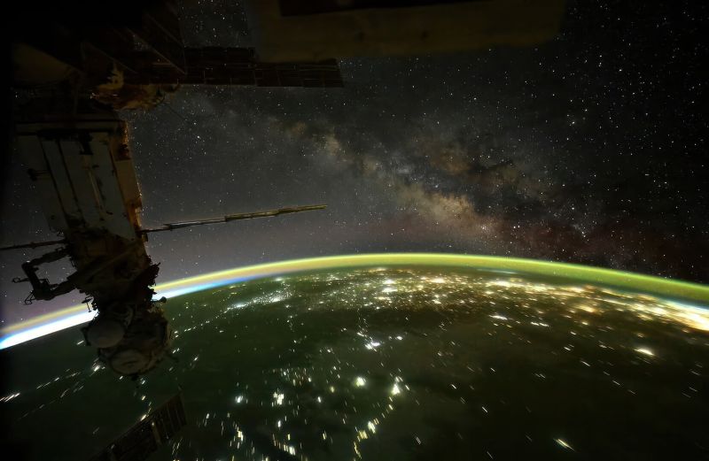 Strut of the space station in silhouette, dark Earth below with city lights, and distant cloudy band of the Milky Way.