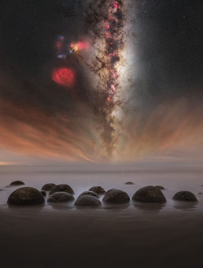 Rounded stones in a lake with the vertical Milky Way above and patches of red nebulosity.
