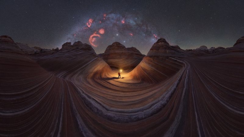 Someone stands with a light at center of a swirling rock formation with the Milky Way arch behind.