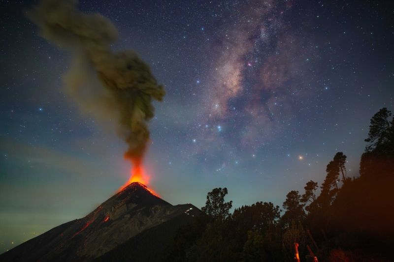 A volcano erupting with red fire and smoke and the Milky Way above it to the right.