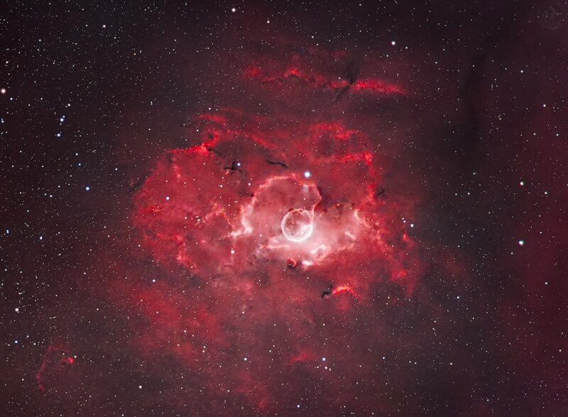 A bright red, bubble-shaped cloud, surrounded by red swirls of gas over a multitude of stars.