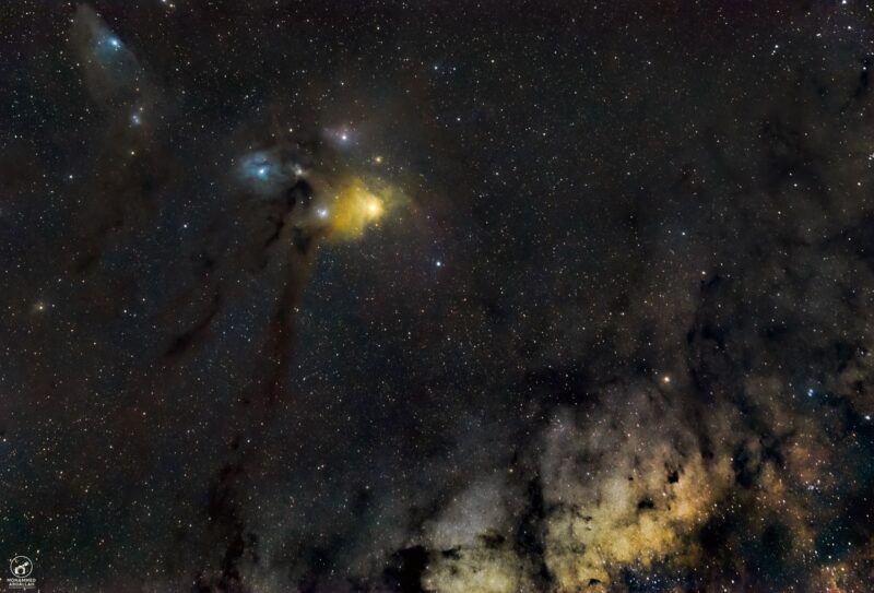 Three patches of reddish, bluish and yellowish nebulosity with dark lanes and a multitude of stars.