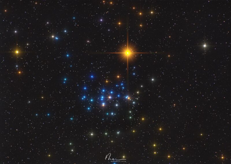 A large grouping of colorful stars with a nearby bright orange star-like body.