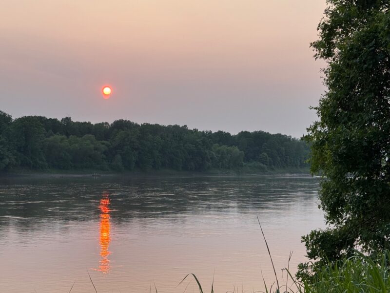 A red sun shining through thick smoke and reflected in a body of water.