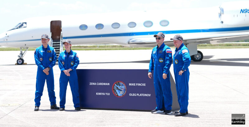 Crew-11 mission: 4 people in blue uniforms stand by a sign with their names on it in front of an airplane.