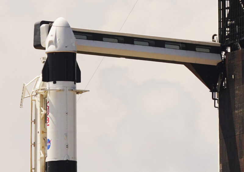 A capsule atop a standing rocket with an arm extending out to it from a gantry.