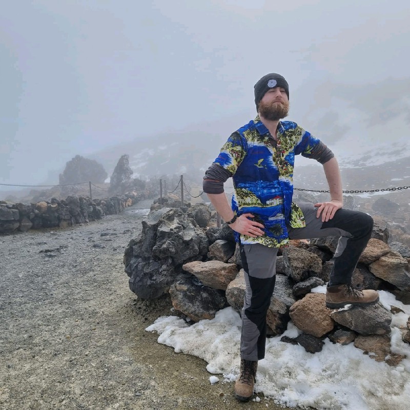 Man with beard posing on a rock wall with some snow on the ground, in a misty landscape.