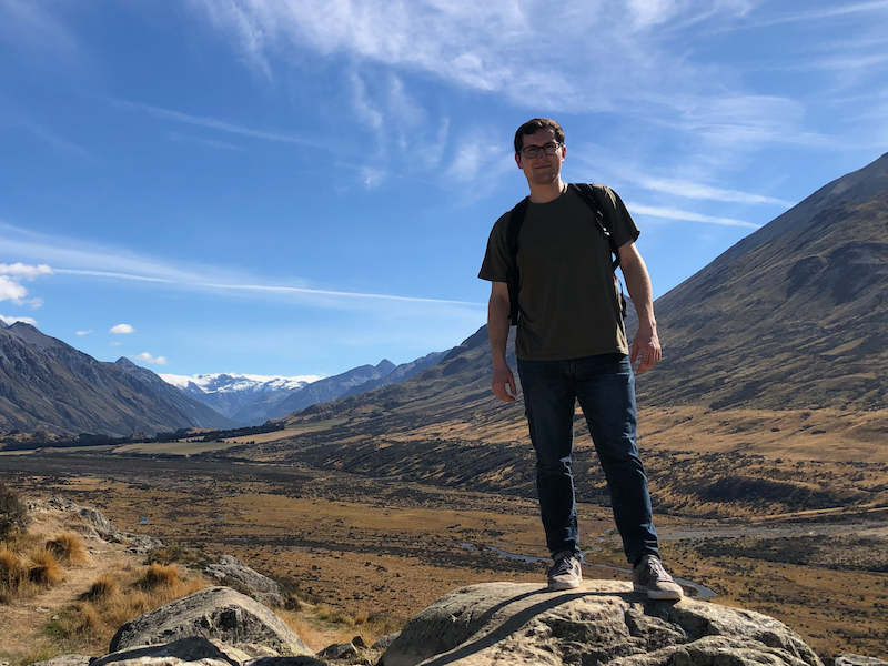 Man in t-shirt and jeans standing on a large rock with a valley and blue sky with wispy clouds behind him.