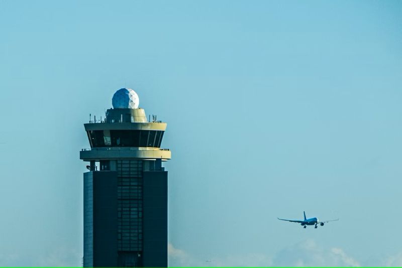Blue sky with an airport control tower and a radar dome on top with a plane to the right.