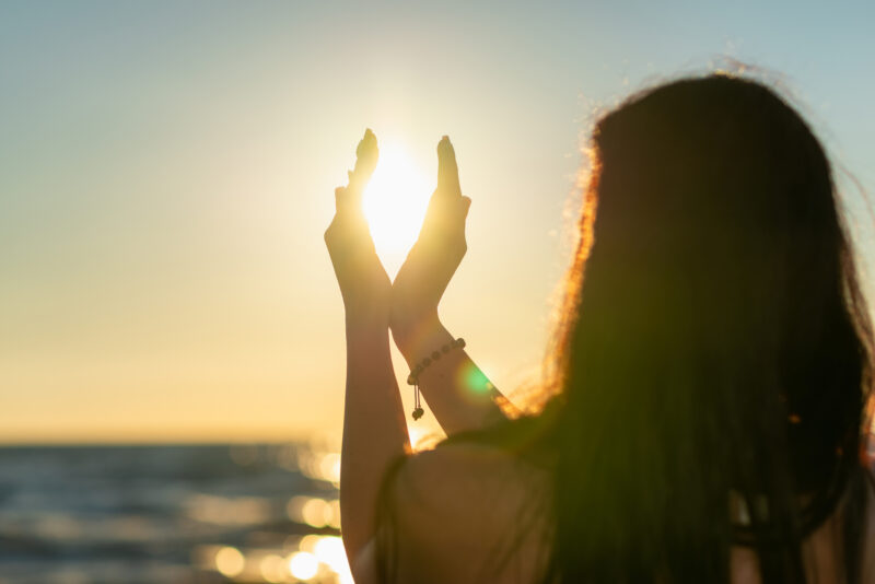 Silhouette of a young woman, her hands cupping the low sun, against the background of the sea.