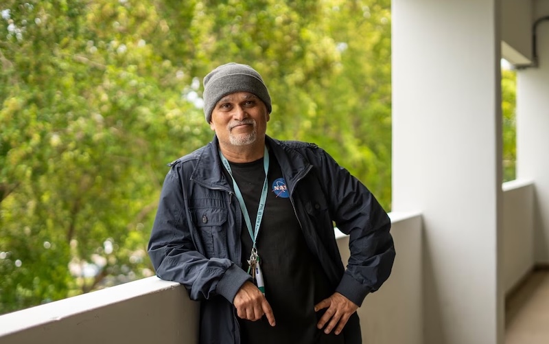 Man wearing a knitted cap, jacket and lanyard leaning against a low wall with trees in the background.