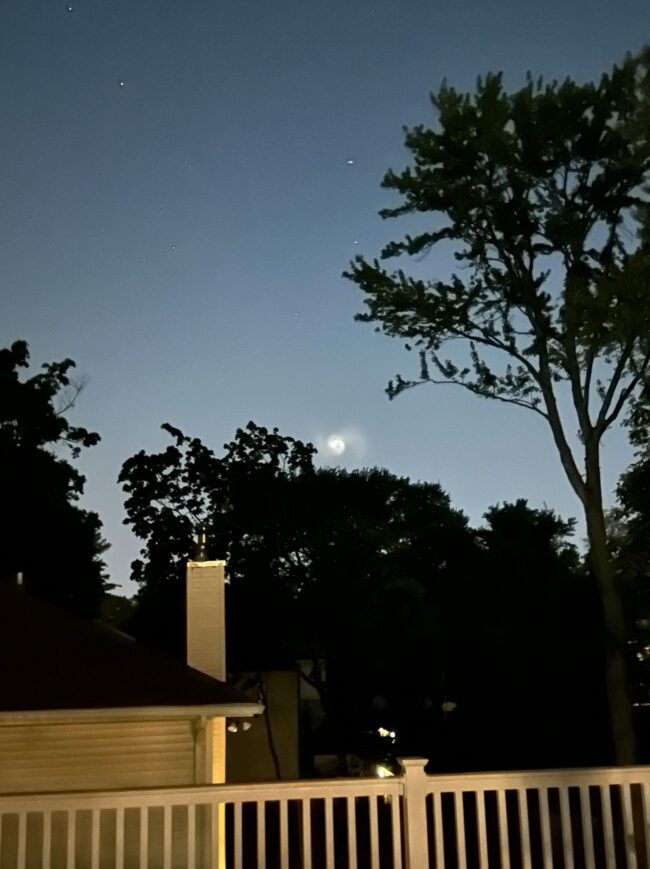 View of trees and a rooftop and chimney in the night with a white spiral just above the trees.