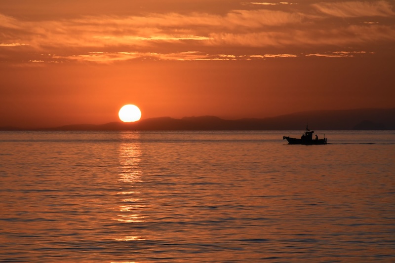 Orangish sky and water with the sun rising in dark twilight over a body of water. A small boat is in the foreground.