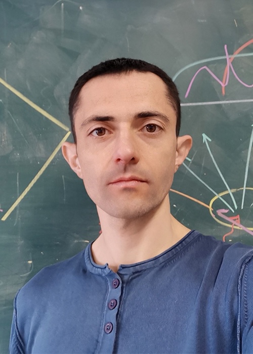 Determined-looking young man with short hair wearing a blue shirt, with a chalkboard behind him.
