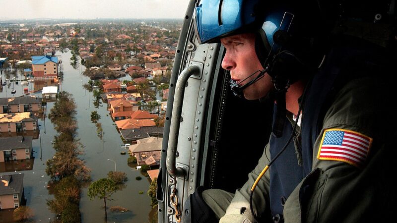 A soldier looks out an open helicopter door, in flight high above a flooded neighborhood.