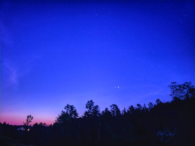 Two bright dots in a blue sky. The horizon looks pinkish. There are trees in the foreground.