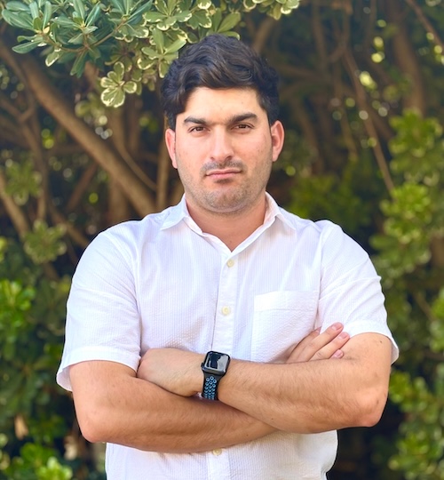 Man with dark hair and wearing a white shirt standing with his arms folded. Trees are in the background.